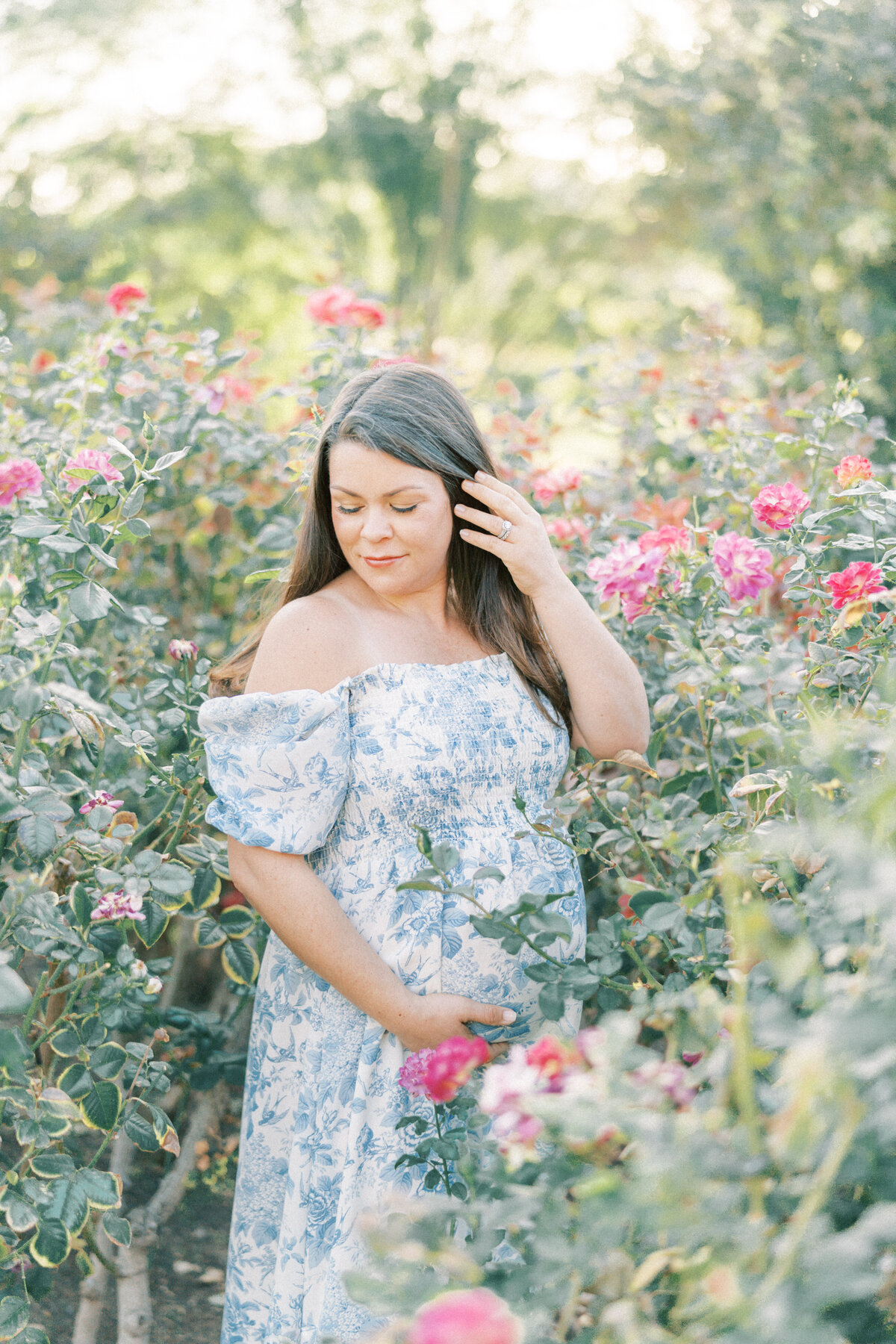 pregnant mom in blue and white dress holding her belly and hair back surrounded by roses
