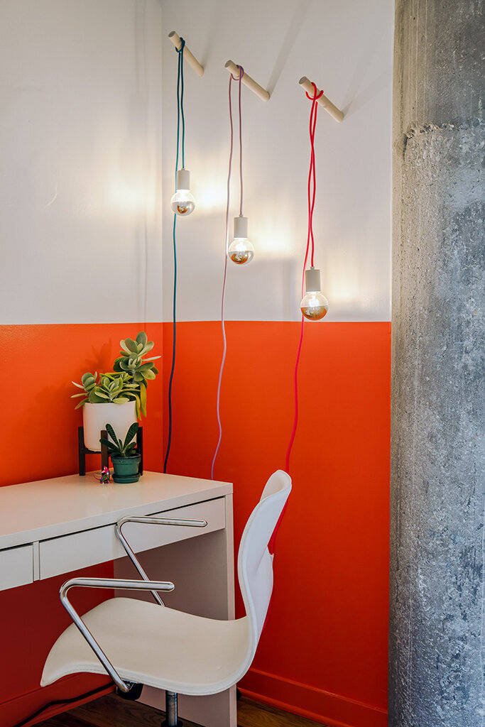 A modern white desk sits against a wall that is half white and half vibrant orange. Three pendant lights with exposed bulbs hang from the top of the wall on colorful cords (teal, pink, and red), each attached to a small wooden peg. A white chair with chrome legs is positioned in front of the desk. A small white planter with a succulent and another tiny plant sits on the desk.