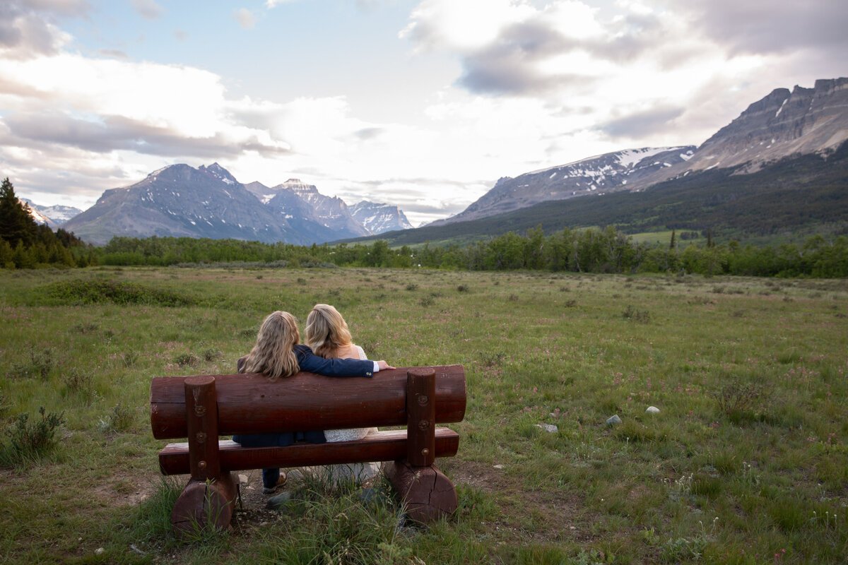 Two brides sit on a bench in Glacier National Park watching the sunset behind the mountains on their elopement day.