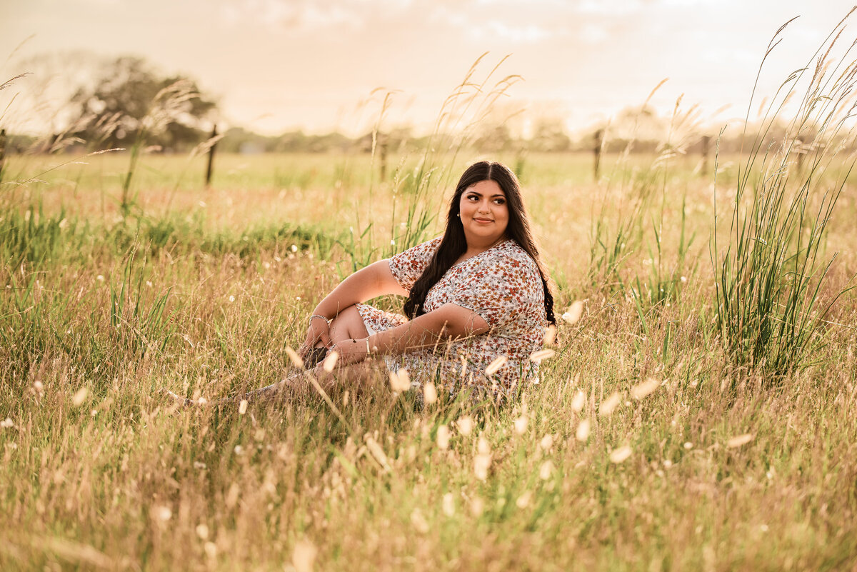 A senior girl sits in a field of long golden grass at sunset in Santa Fe, TX.