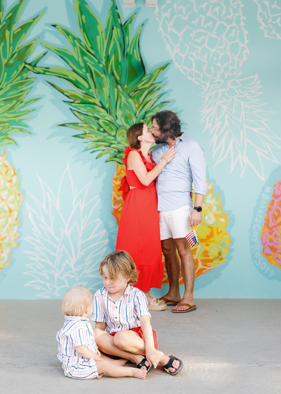 A couple shares a kiss in front of a vibrant pineapple mural while their children sit nearby during a Florida Keys family session.