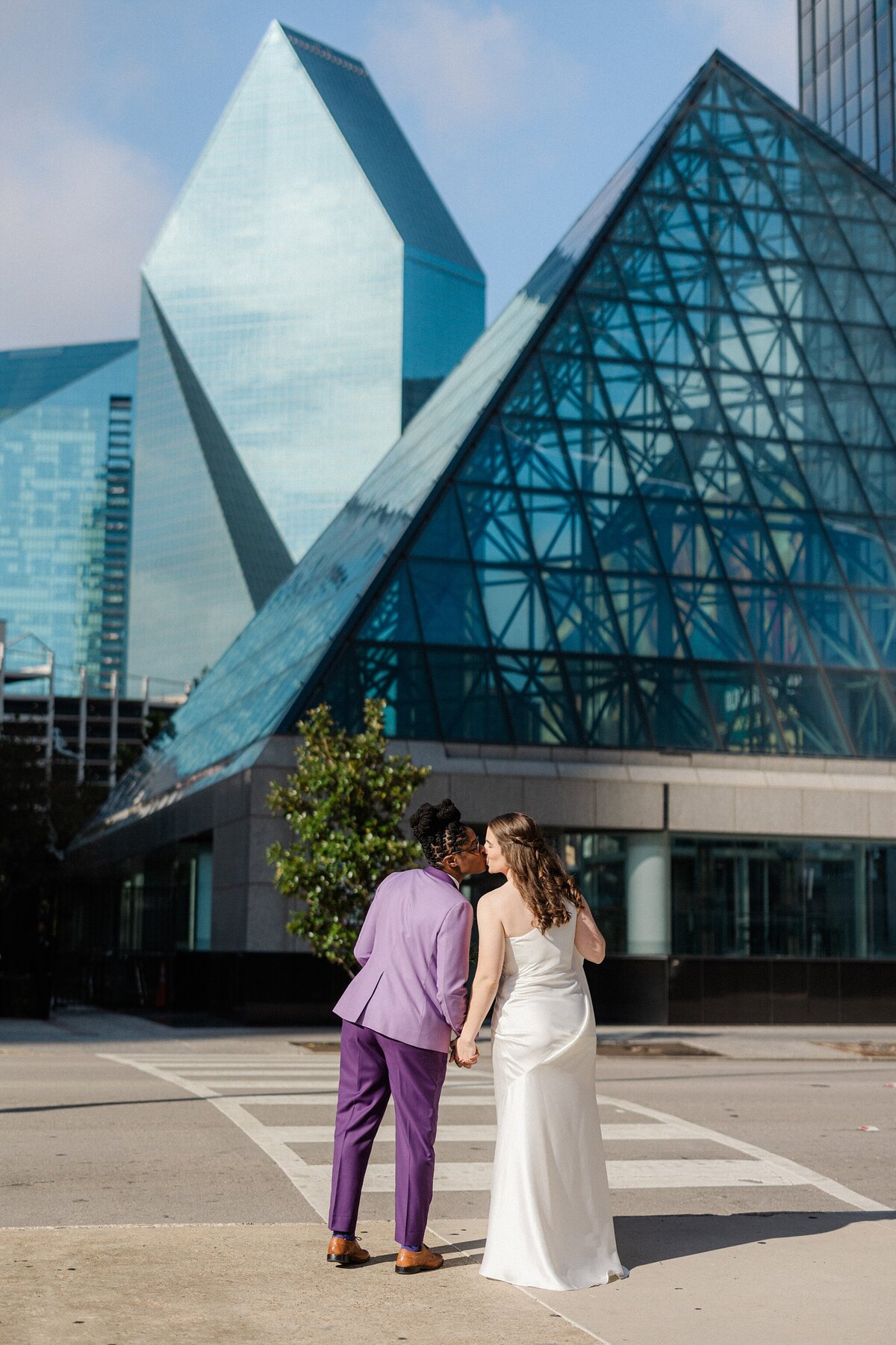 Two brides share a kiss outside of The Westin Dallas Downtown in Dallas, Texas. The bride on the right is wearing a sleeveless, elegant, white dress. The bride on the left is wearing a purple suit with dress shoes. The Dallas skyline can be seen in the background.