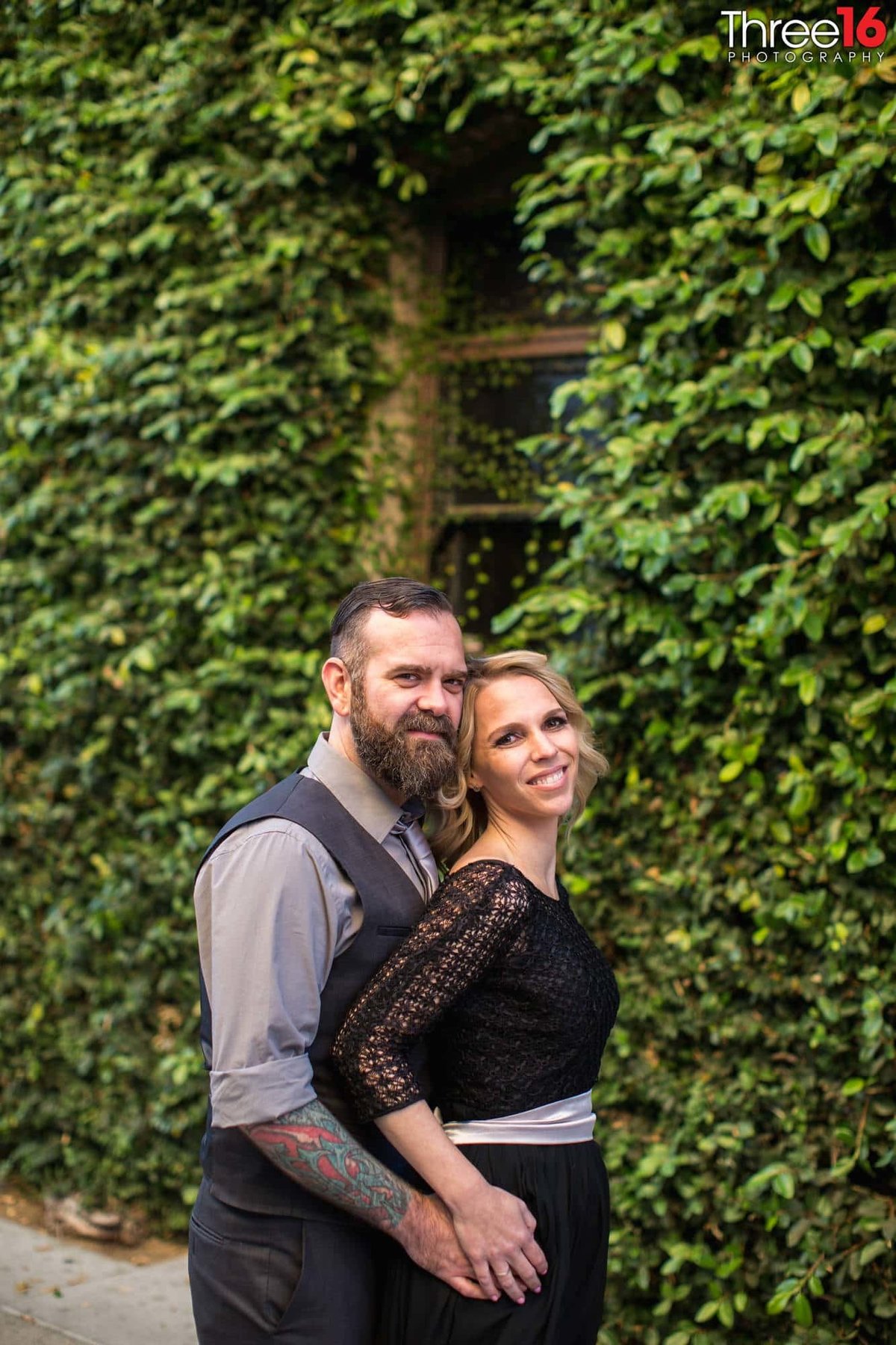 Groom to be snuggles up to his Bride from behind in front of ivy covered wall