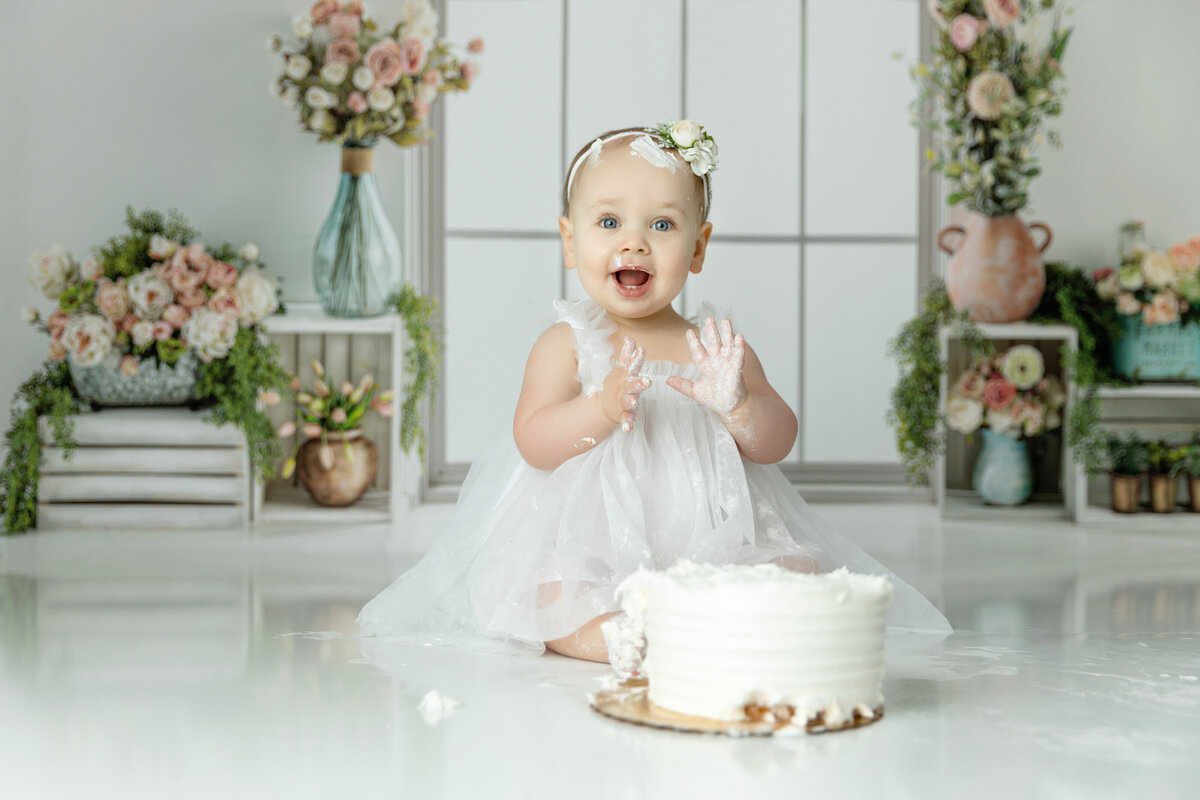 A happy toddler girl in a white dress claps her cake covered hands together in a studio