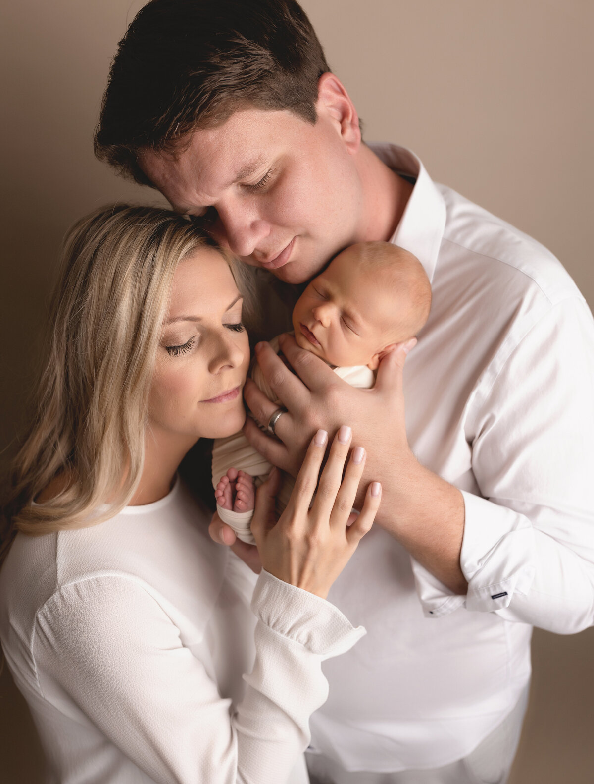Family newborn photo in West Palm Beach and Boca Raton newborn photography studio. Baby is wrapped in a cream knit swaddle. Dad is holding baby's back against his chest, one hand under baby's bum, the other on baby's chest. Mom is facing dad and baby and resting her hand on top of dad's forehand. Mom and Dad have their heads touching and eyes closed.