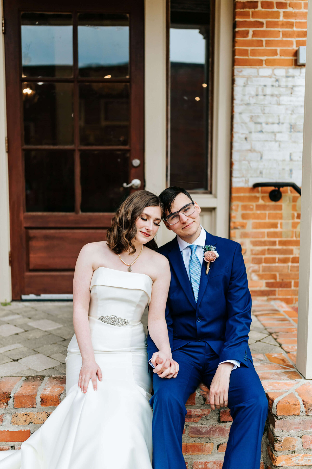 A bride in a white strapless dress and a groom in a blue suit sit on brick steps, holding hands and leaning their heads together, smiling gently in front of a rustic building.