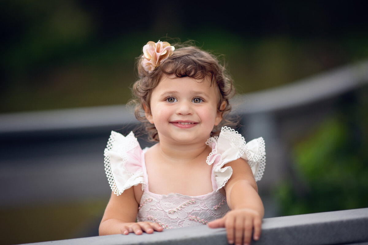 A toddler girl in a pink frilly dress smiles while standing on a park bridge