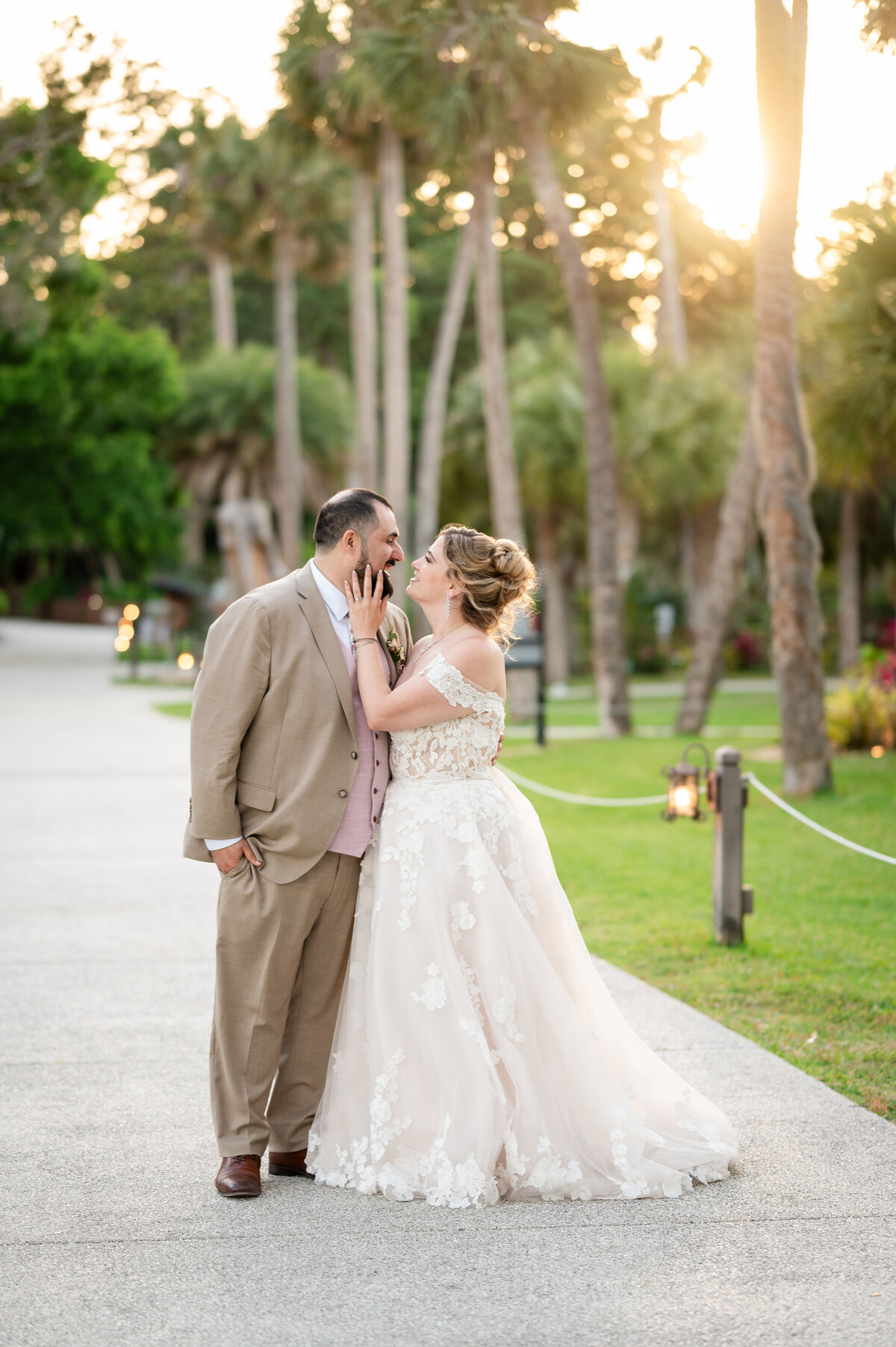 A bride and groom share a tender, joyful moment at golden hour in St. Augustine, Florida. Surrounded by palm trees and soft evening light, the bride gently touches the groom’s face while smiling, her lace gown glowing in the sun. The groom, wearing a tan suit and blush vest, leans in lovingly. Captured by Heartful Films, a Jacksonville wedding photographer and videographer known for romantic, true-to-color storytelling set in beautiful natural backdrops.