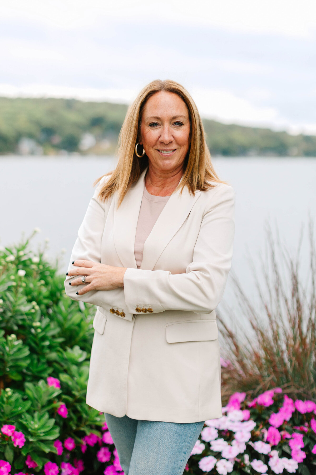 A woman with shoulder-length blonde hair stands with her arms crossed, wearing a cream blazer and gold hoop earrings. She is outdoors near a lake, surrounded by lush greenery and vibrant pink flowers, with a backdrop of distant hills and water