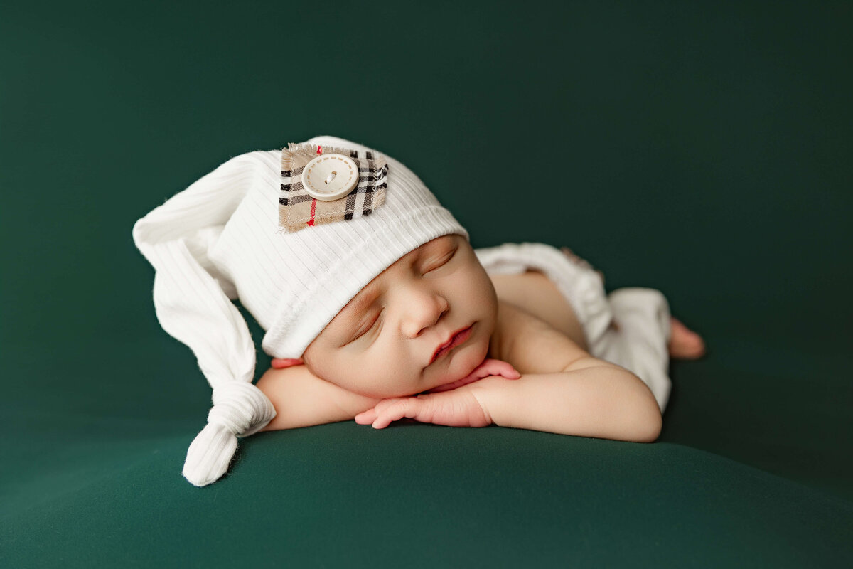 A newborn baby rests peacefully on a dark green surface, wearing a soft white hat adorned with a plaid accent. The baby’s eyes are closed, and they appear comfortable, showcasing a serene expression while sleeping.