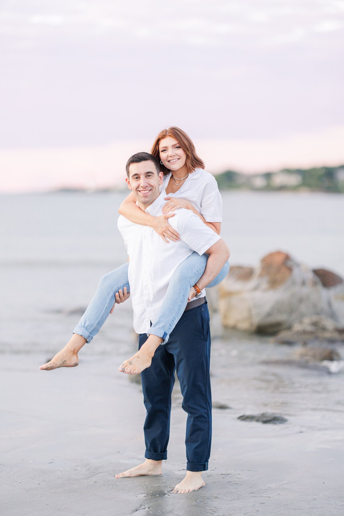 A man and woman smiling at the camera as the man gives the woman a piggyback ride on Wingaersheek Beach representing a Gloucester engagement session.