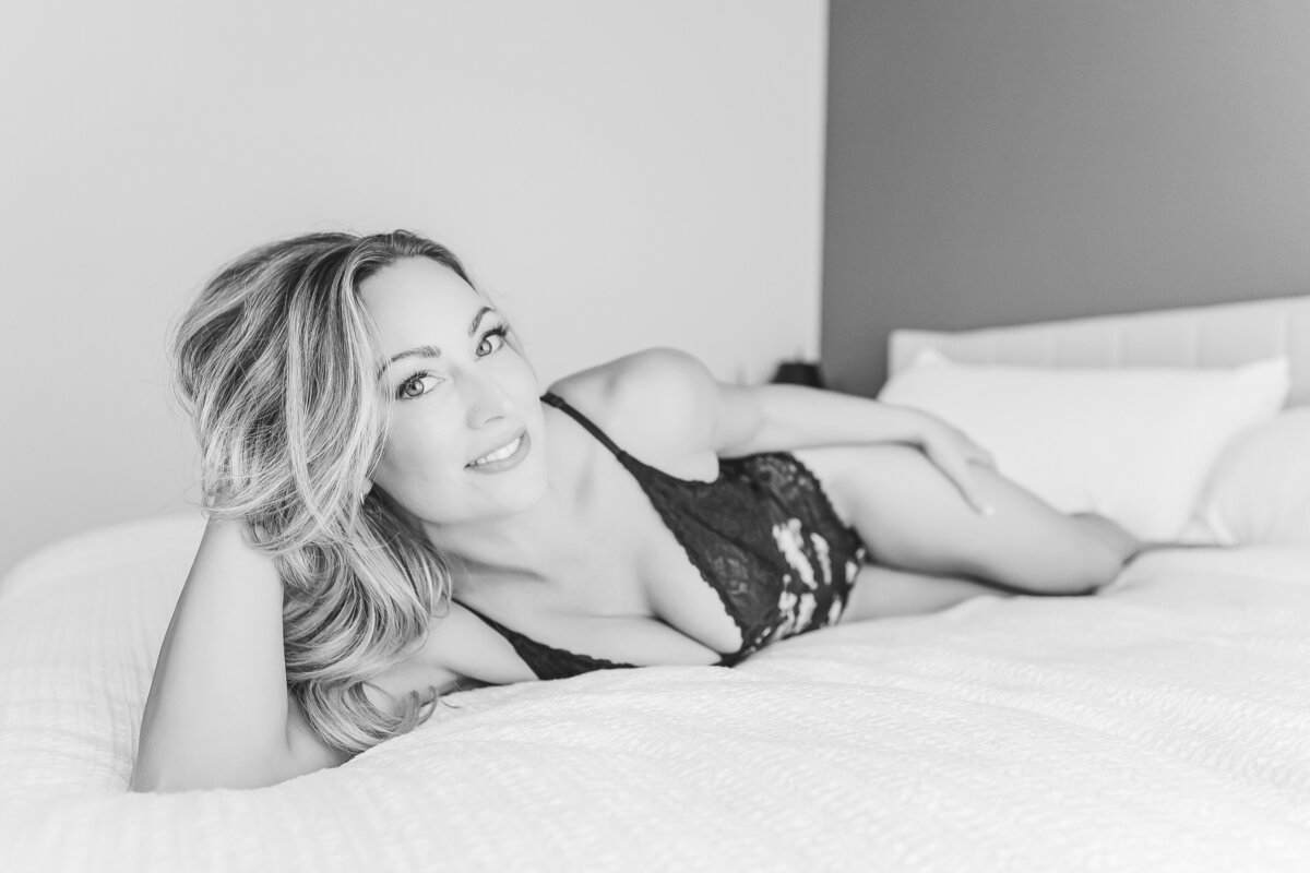 A woman lying on a bed in black lingerie, smiling with a playful and confident look, enjoying the relaxed setting.