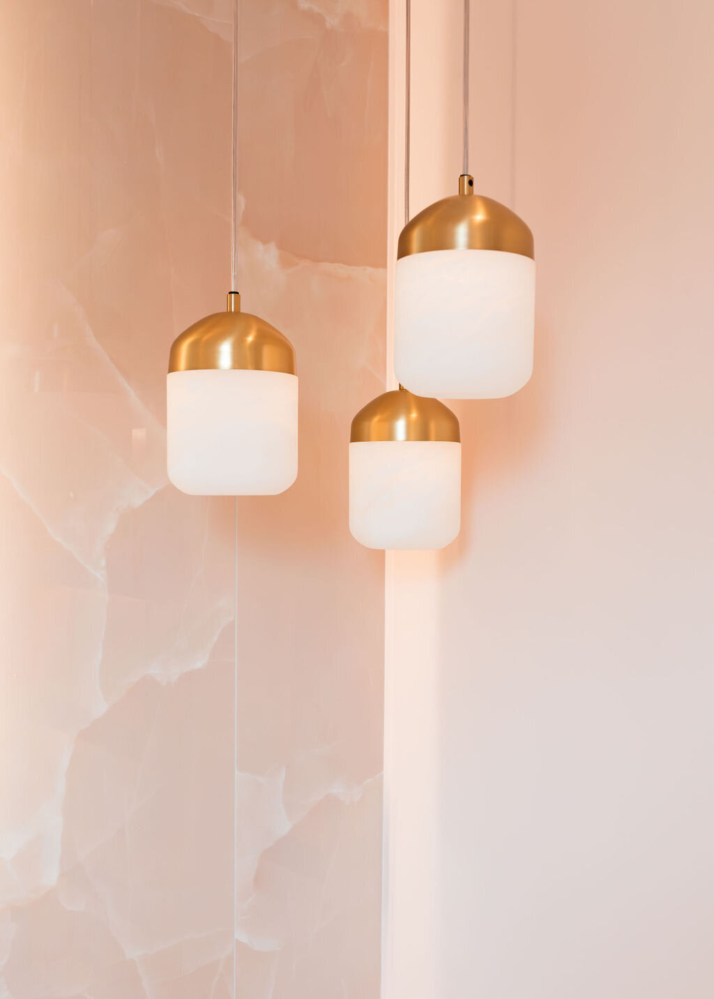 A trio of white and gold pendant lights hanging in the corner of a pink marble bathroom vanity.