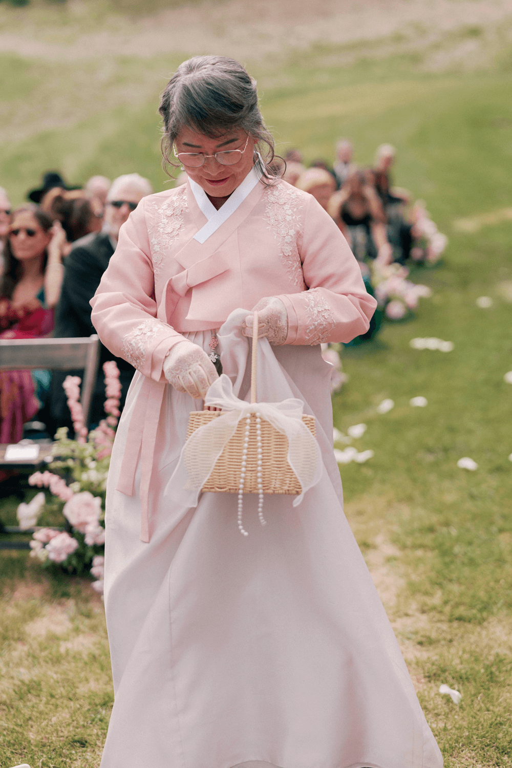 Grandmother serving as flower girl at a pastel summer wedding at Moonlight Basin, wearing a pink hanbok and holding a woven petal basket.