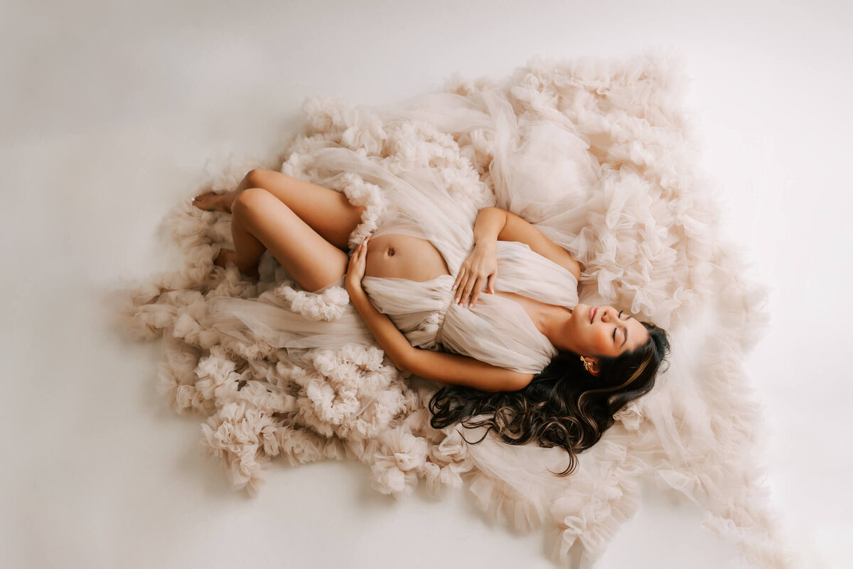 stunning maternity portrait of a brunette mom laying in couture tulle dress holding belly in portland oregon studio.