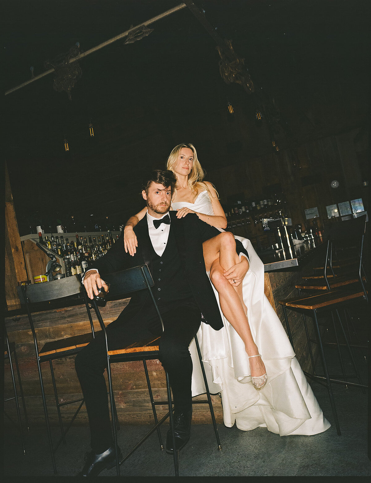 A man in a tuxedo and a woman in a white dress seated at a bar, with the woman draping her leg over the man's. The bar background is dimly lit with bottles