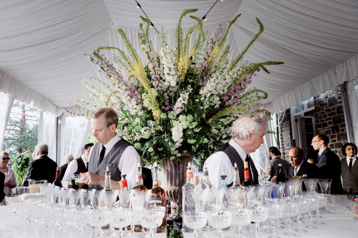 A table with many wine glasses and drinks, with two men and a large flower arrangement behind it.
