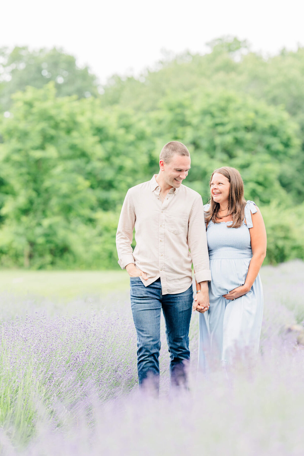 Pregnant woman in a light blue dress laughs with her husband while walking through a lavender field