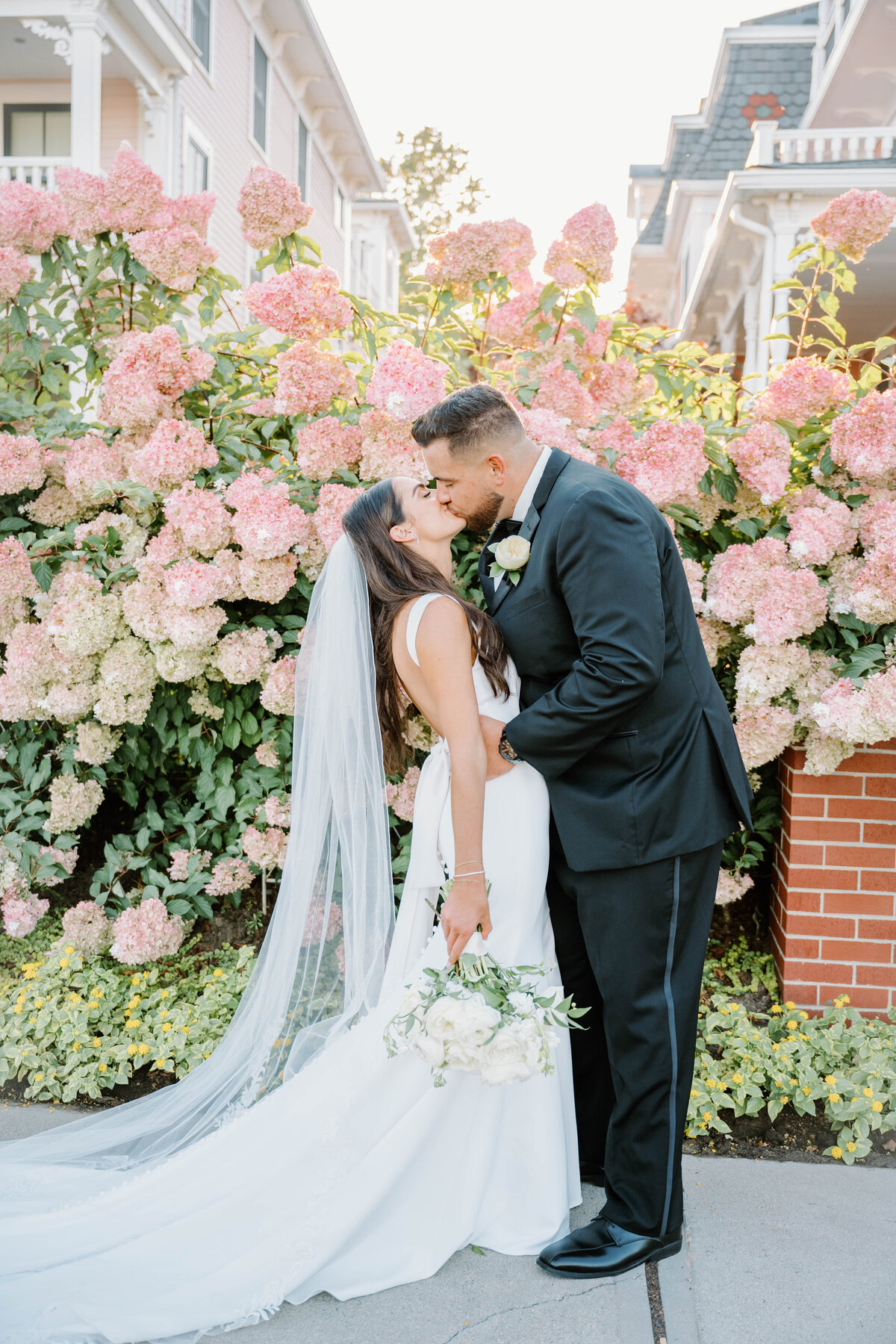 Rhode Island Wedding Photographers | A bride in a white gown and veil kisses a groom in a black suit before a lush background of blooming pink hydrangeas. She holds a bouquet.