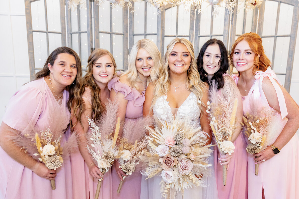 The bride poses with her bridesmaids, all holding dried floral bouquets in warm, neutral hues.
