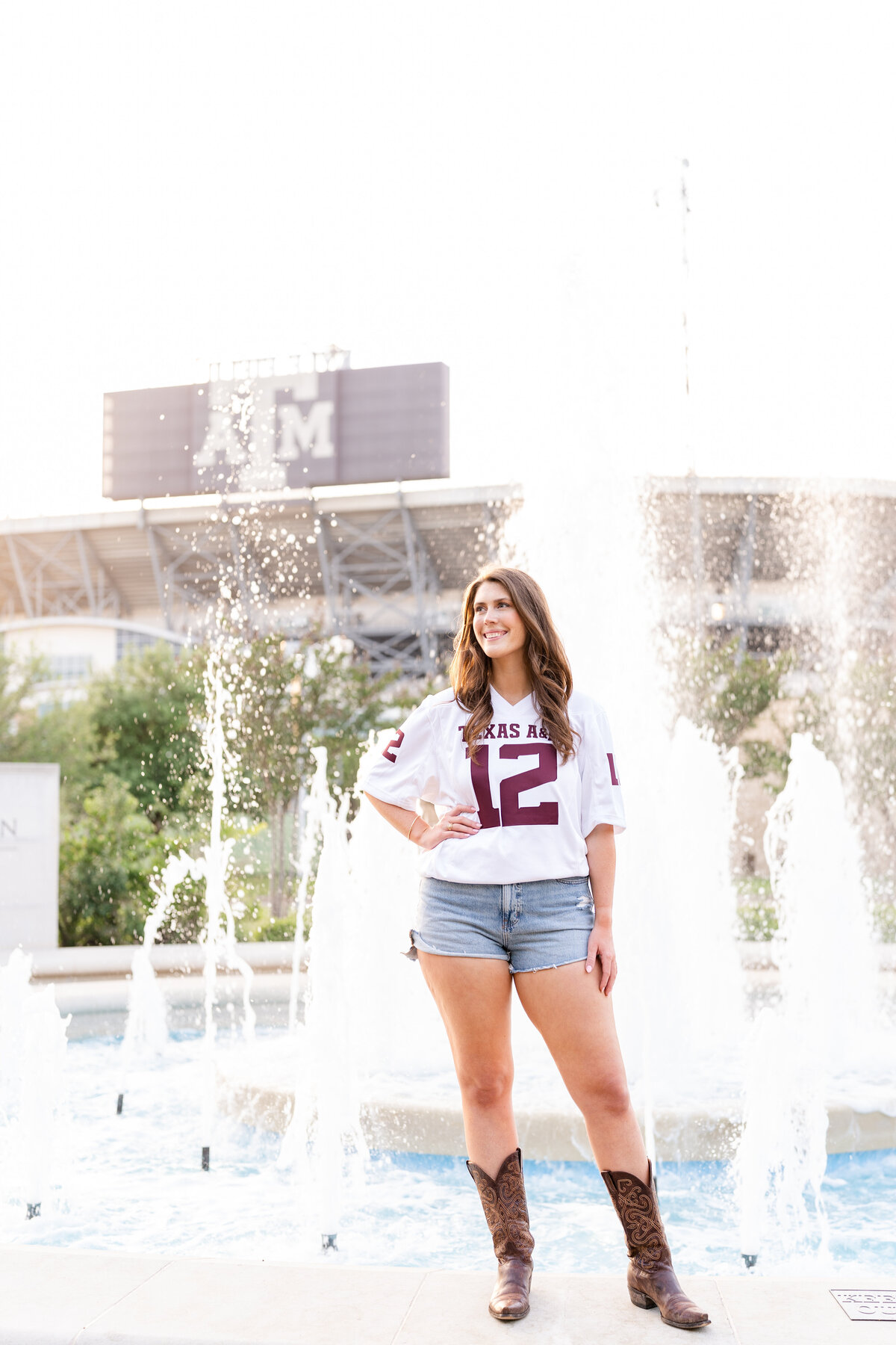 Texas A&M senior girl standing on ledge of fountain with hand on hip and wearing white Aggie jersey and cowboy boots with Kyle Field in background