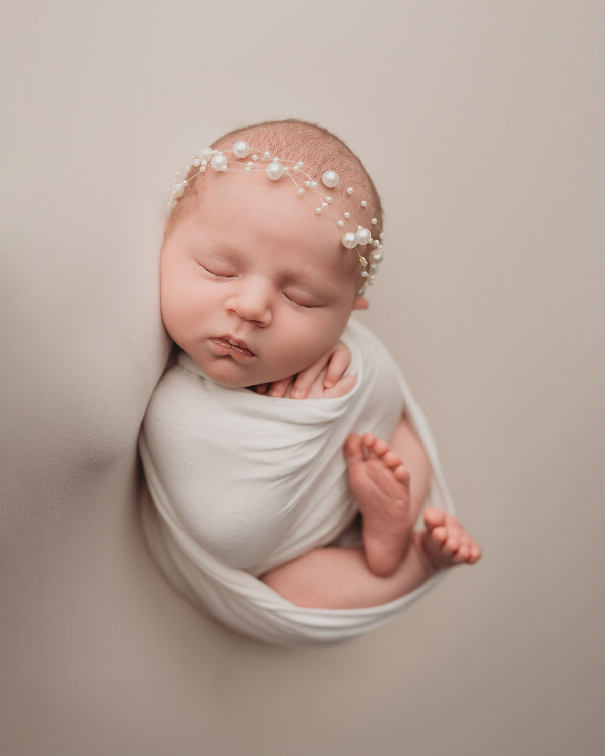 Newborn baby girl wrapped in white swaddle with feet hanging out wearing pearl headband laying on white backdrop