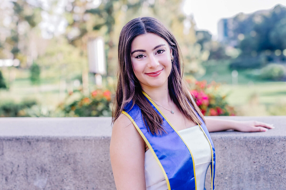 A young woman smiles while wearing a blue and gold sash, standing outdoors. She has long, brown hair and is positioned against a concrete ledge. A blurry green landscape and colorful flowers are visible in the background.