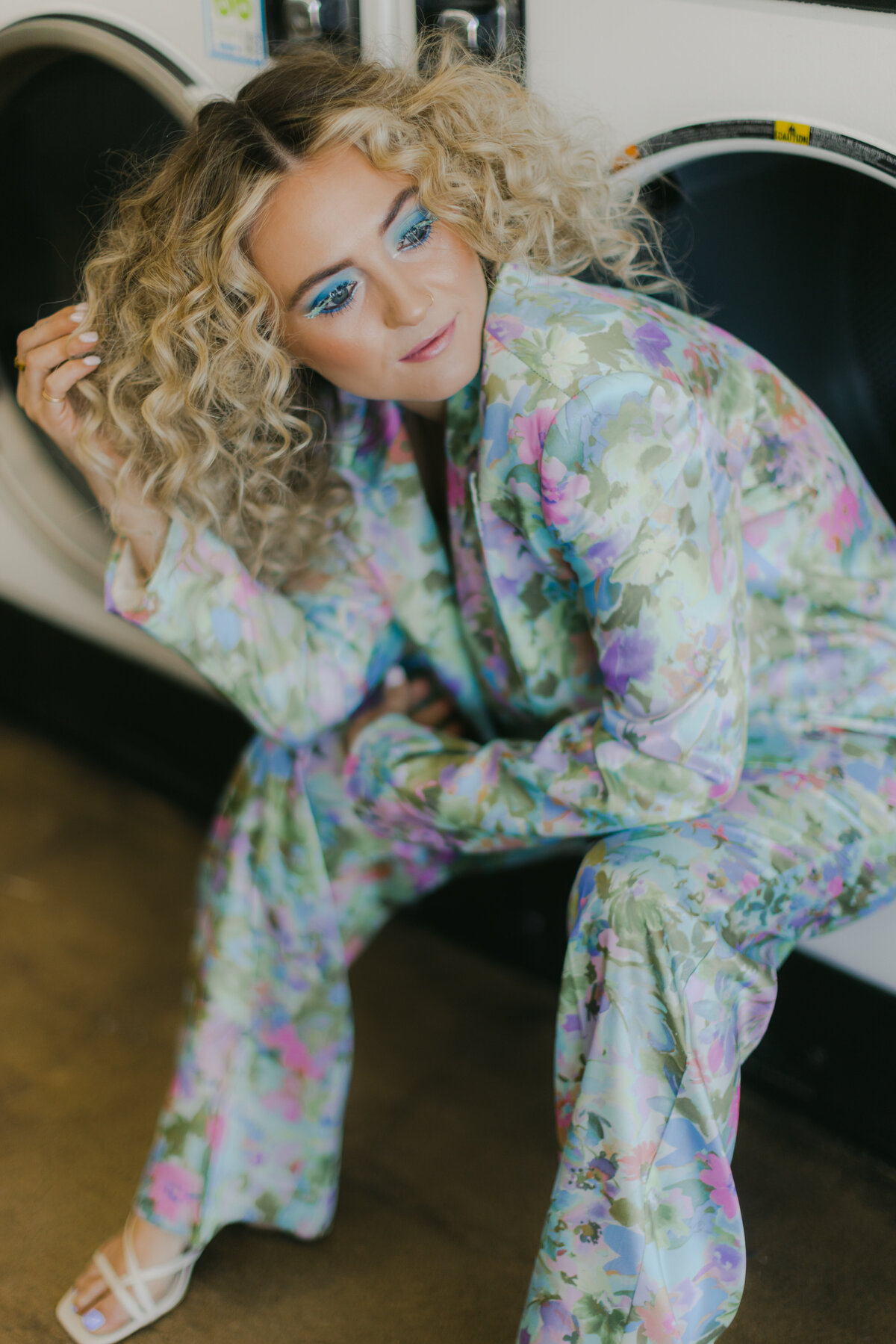 A stylish woman with voluminous curly blonde hair and vibrant blue eye makeup poses confidently in a pastel floral satin suit while crouched in front of a laundromat machine. Captured by Dani Lacey in Greenville SC, this bold branding image highlights vibrant fashion and fearless attitude.