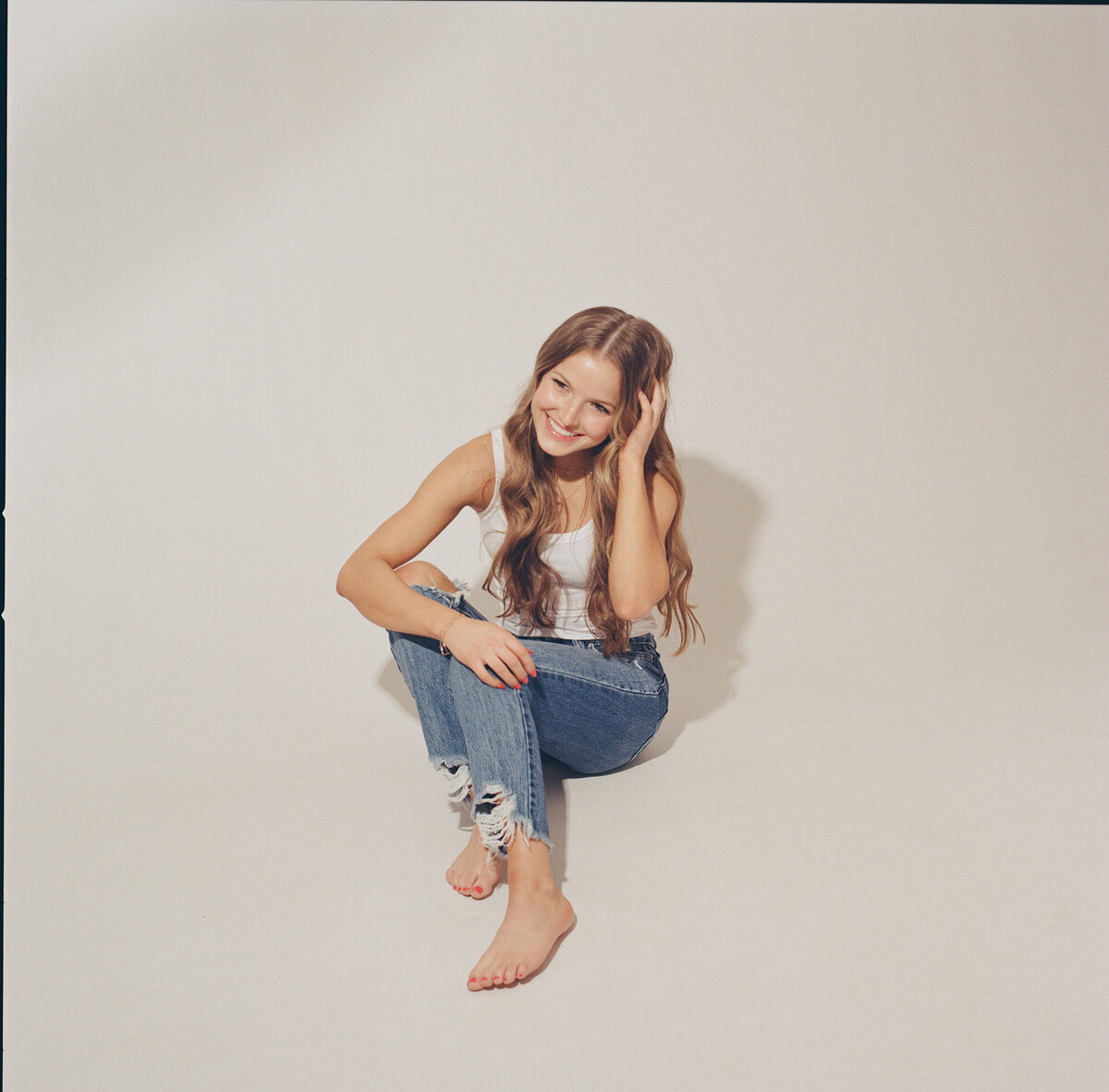 A high school senior sits in jeans and a white shirt with a hand in her hair