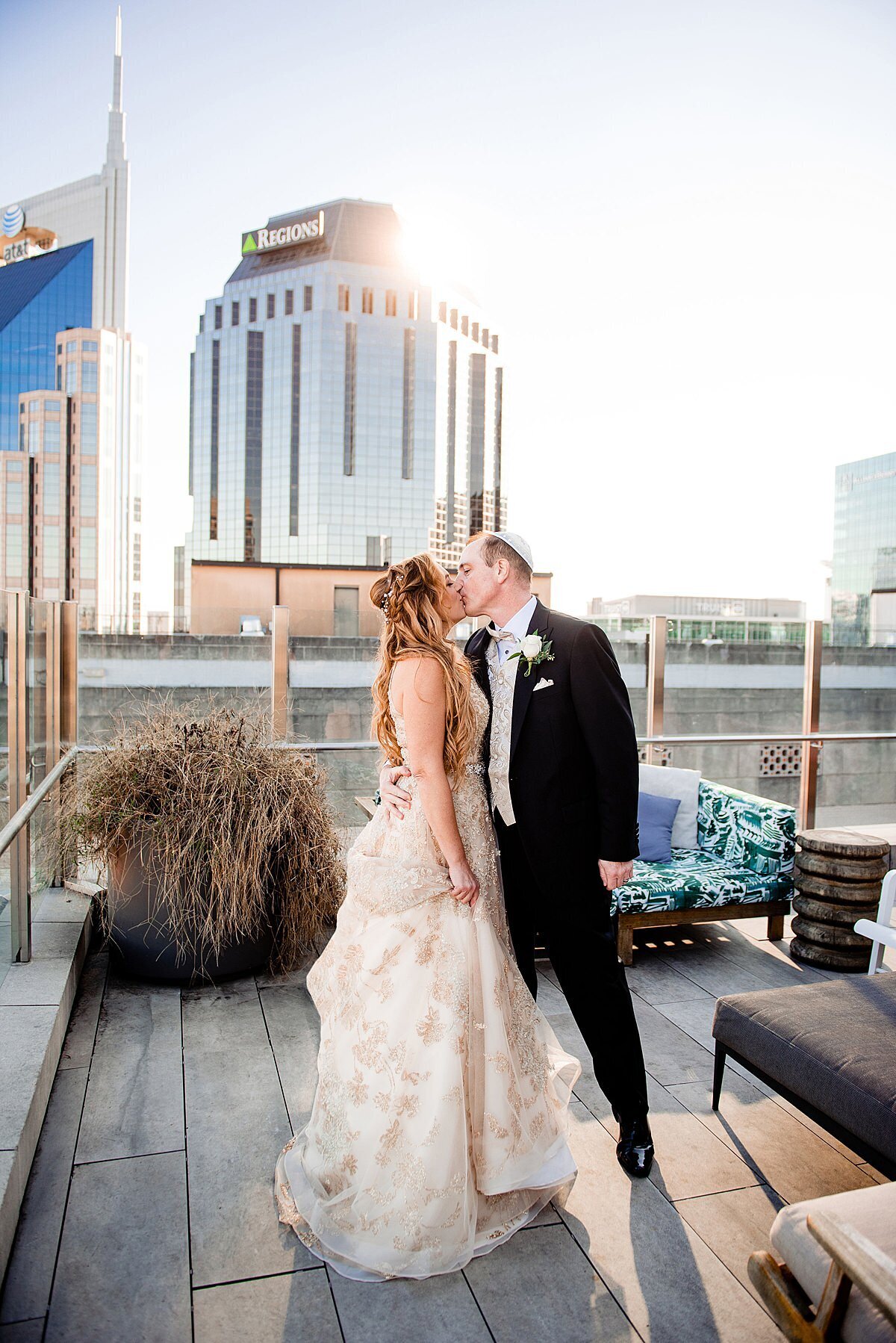 Jewish bride wearing a gold beaded backless gown kisses her Jewish groom wearing a black tuxedo and white yarmulke on the rooftop of the Noelle Hotel in Nashville with the Nashville Skyline behind them.