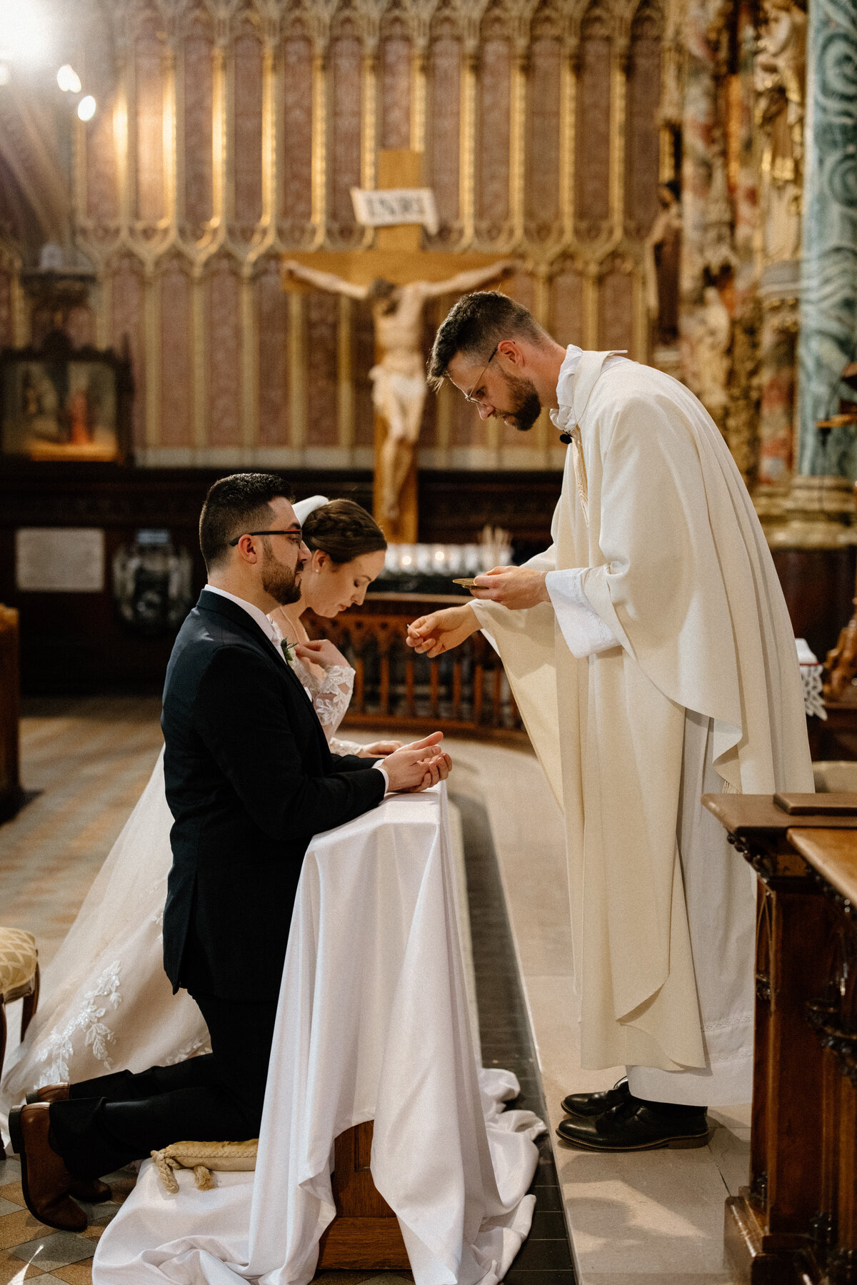 The bride and groom kneels down in front of the priest at the altar during the wedding ceremony