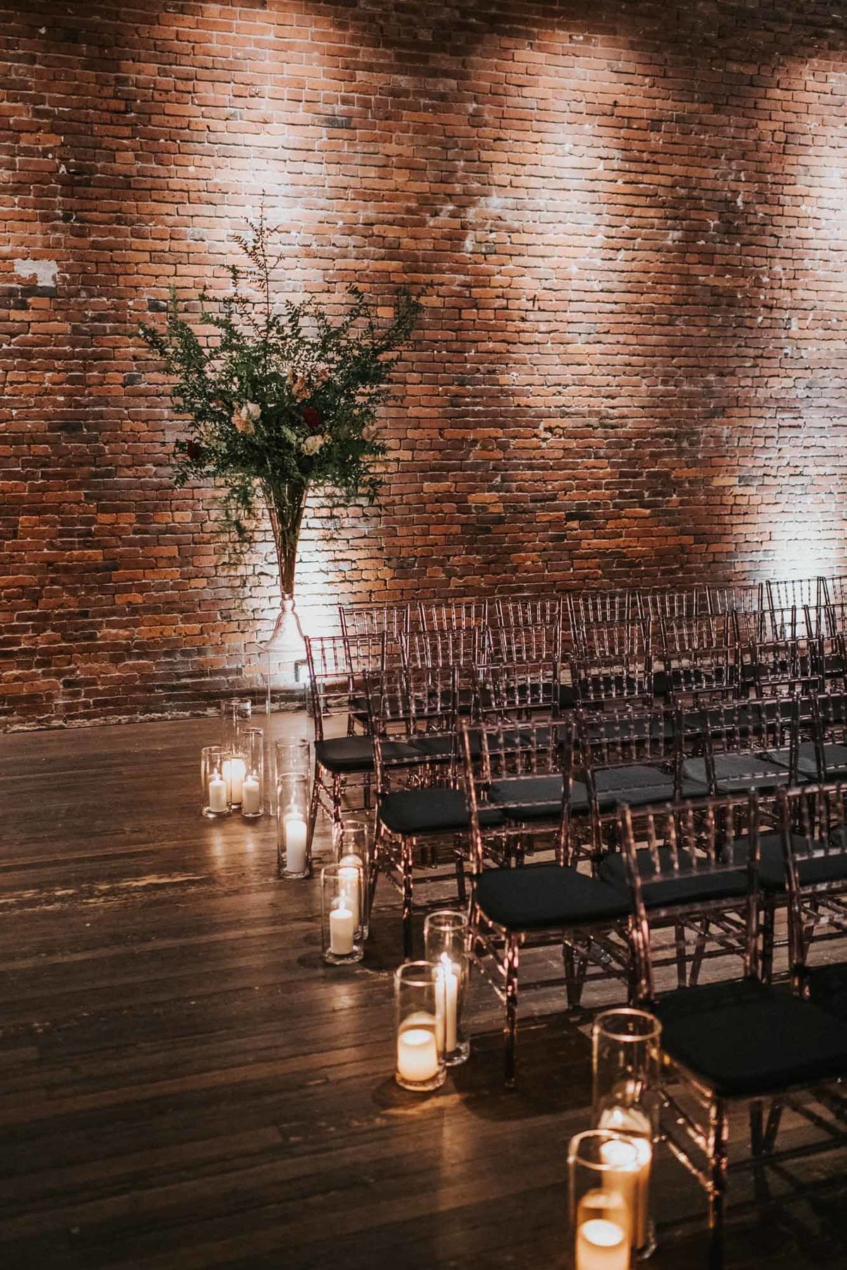 winter wedding ceremony aisle with large greenery bouquet, hurricane candles, and a lit up brick wall in back