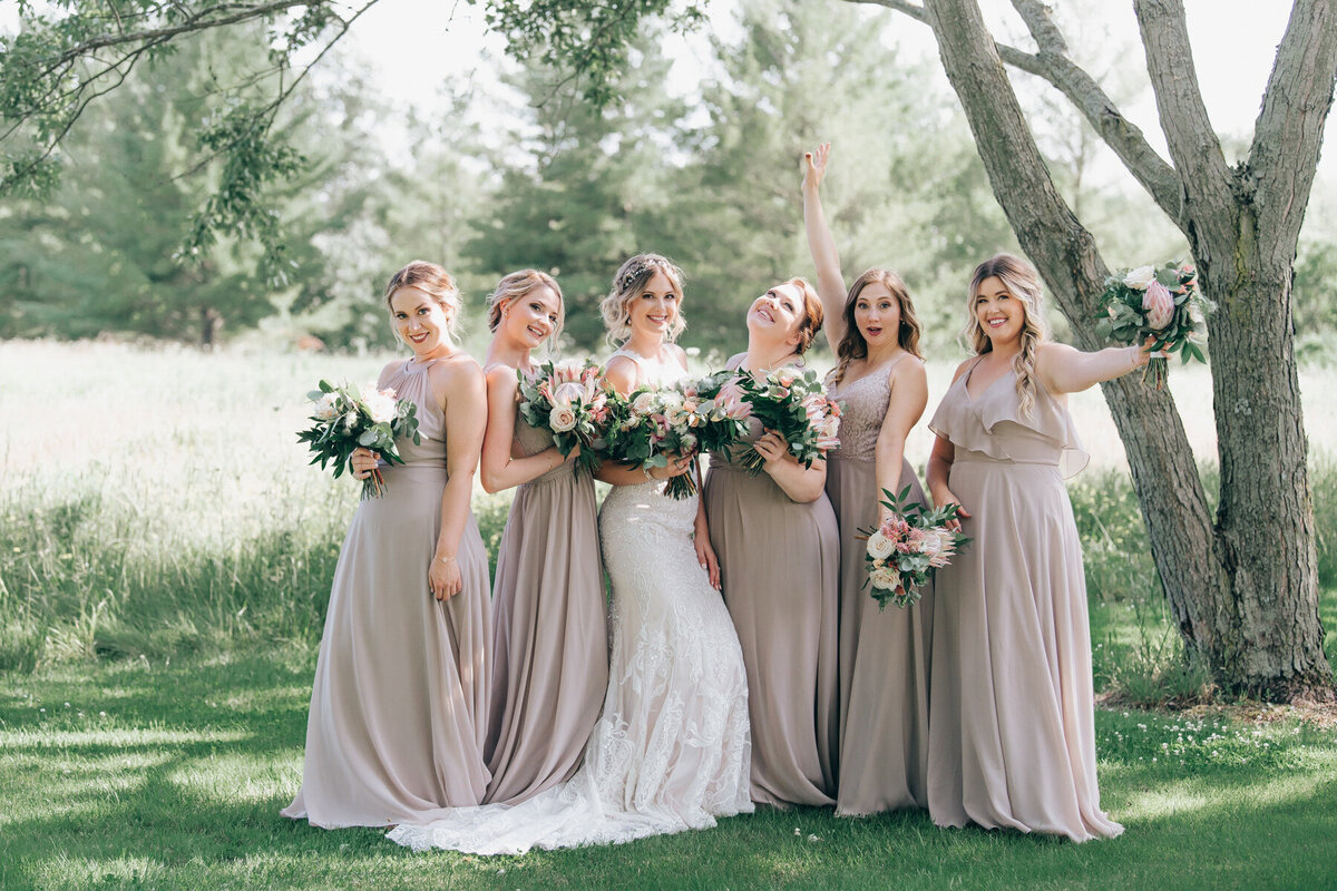 Bridesmaids wearing taupe dresses posing for fun photos with the bride while all holding whimsical pink, ivory and green bouquets