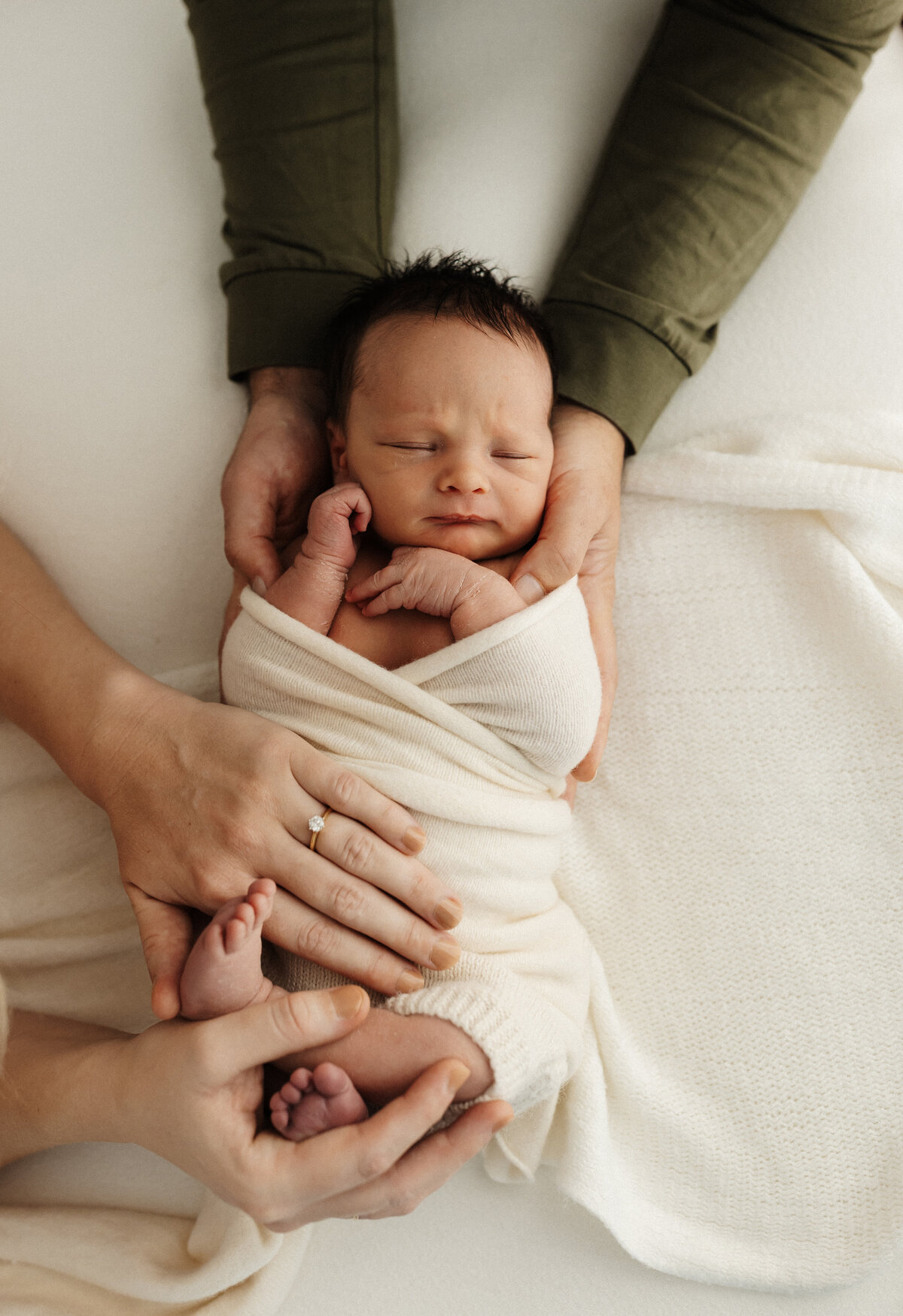 A newborn baby wrapped in a white blanket with his mum and dads hands holding her while she sleeps during their newborn photoshoot in christchurch with lucy jane photography