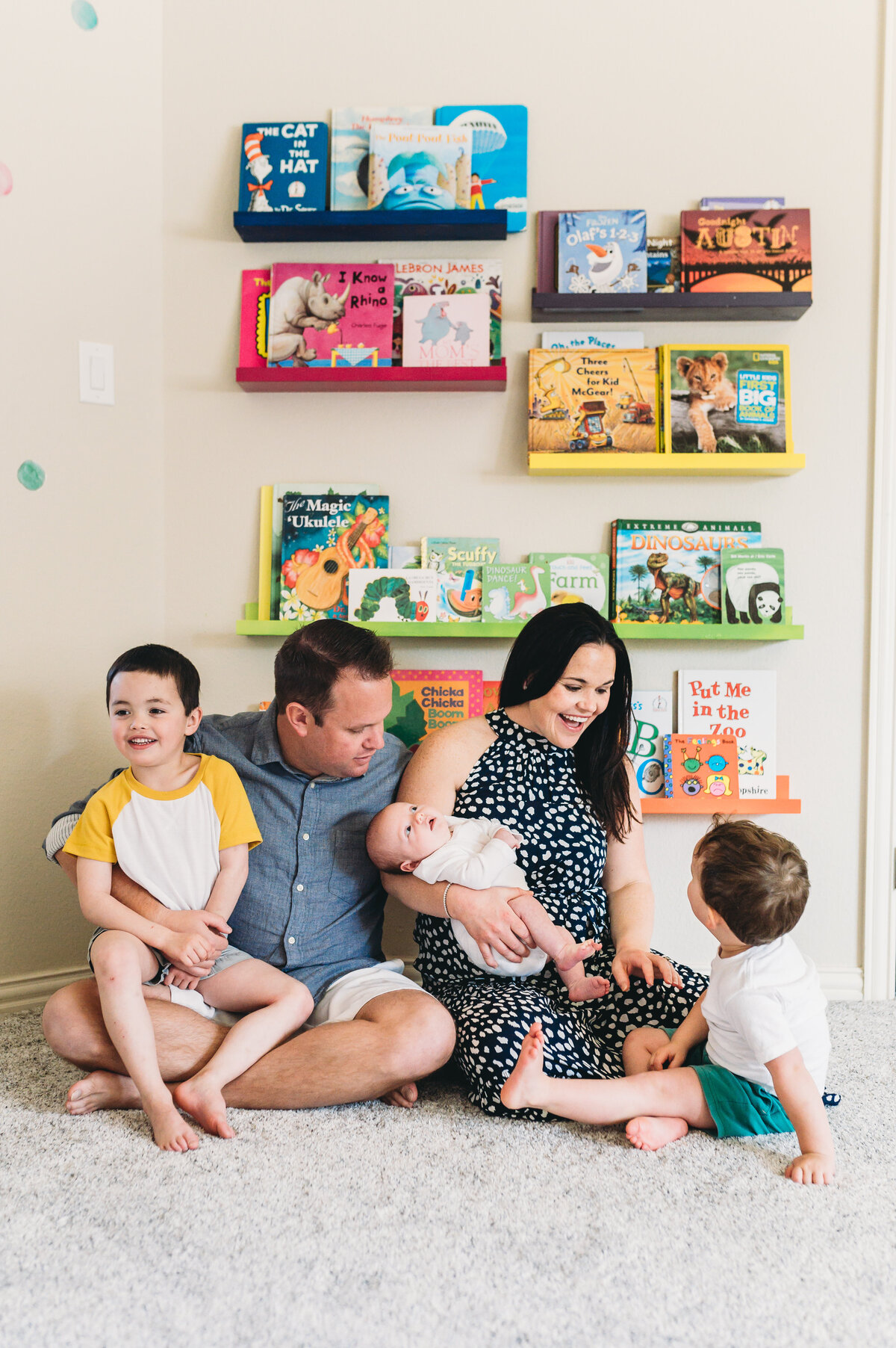 family of five sits on floor at home beneath rainbow book shelves while holding new baby