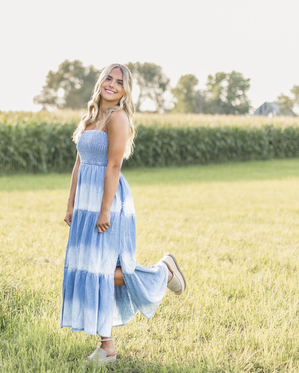 High school senior female with long blonde hair, wearing a blue and white long dress, standing in the grass, with a corn field in the background, pinching the sides of her dress, looking at the camera with one foot up and smiling