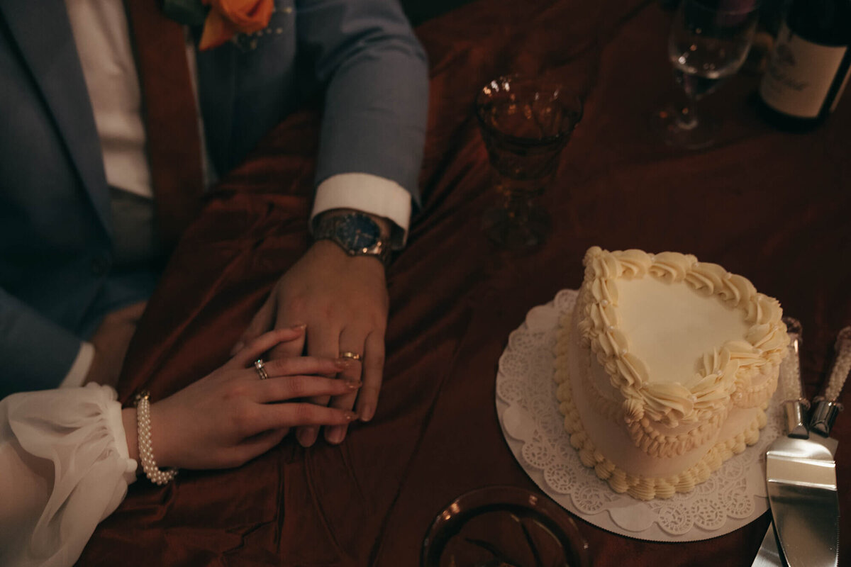 A bride and groom holds hands resting on a table next to a heart shaped wedding cake.
