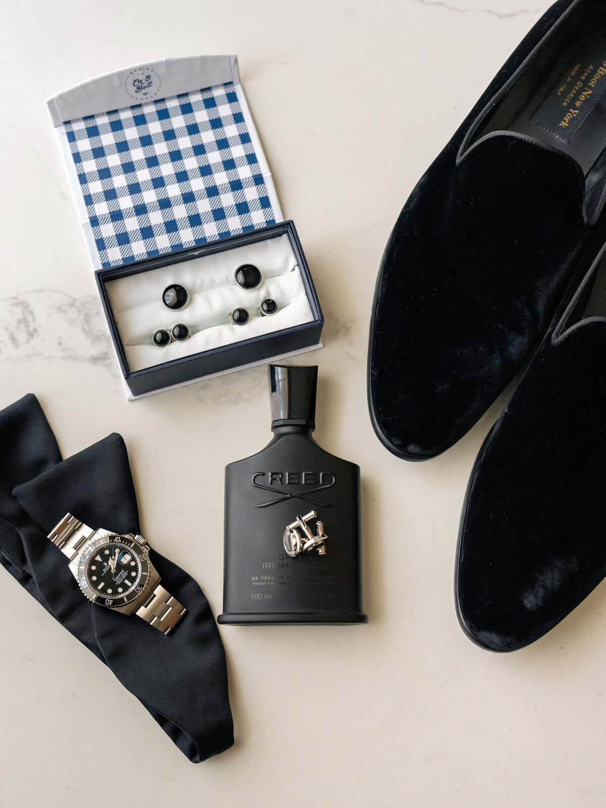 Mens accessories arranged on a marble surface: a box with cufflinks, black formal loafers, a Creed cologne bottle, a black bow tie, and a luxury watch.
