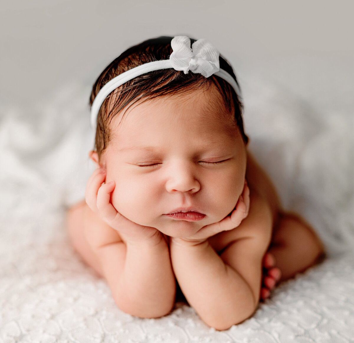 Newborn baby girl on a white backdrop posed in the froggy pose with hands on her cheeks wearing a white bow