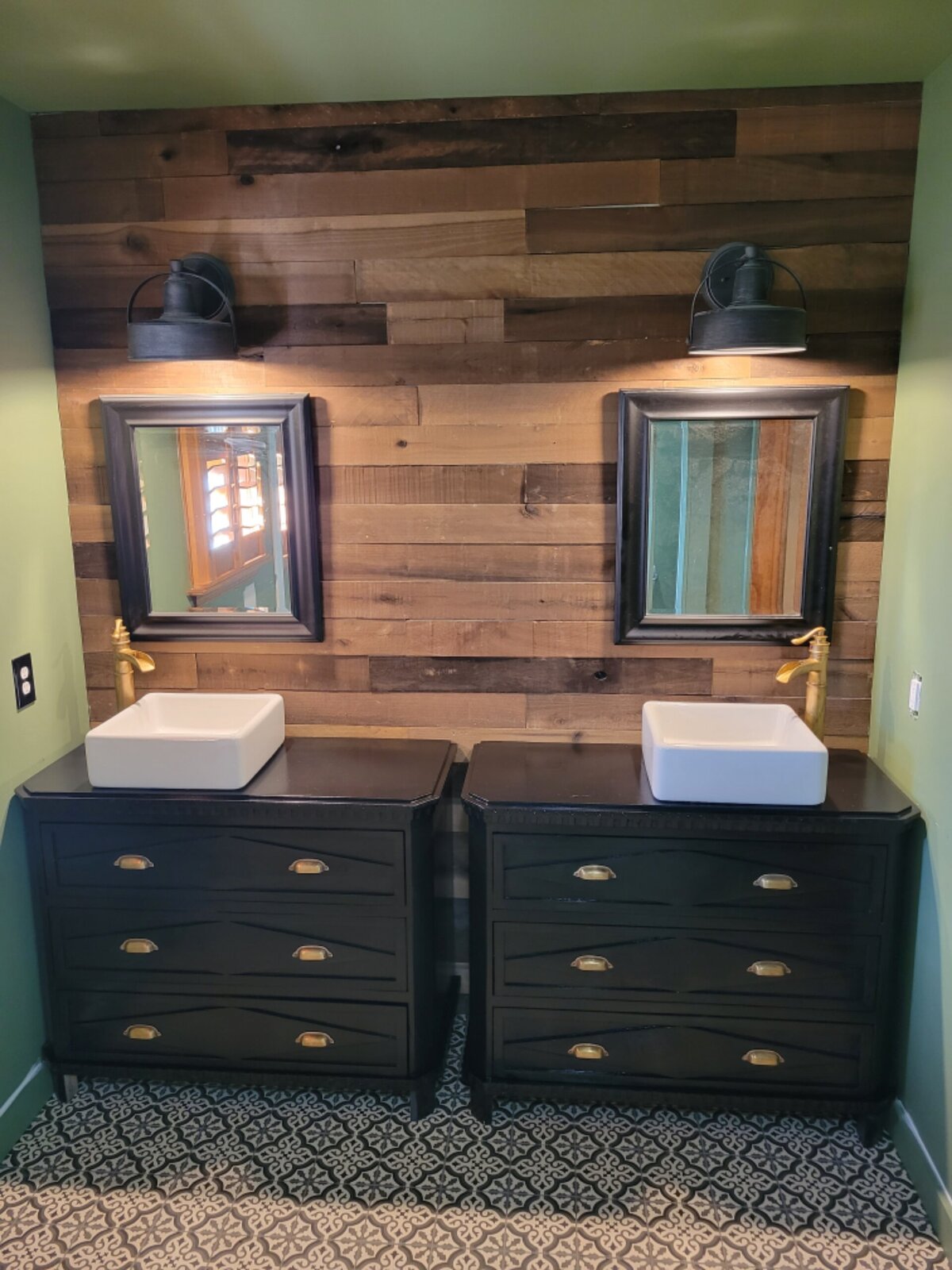 Wooden wall, with black painted cabinets and brass  cup handles with white sink vessels in a green bathroom with patterned tile floor