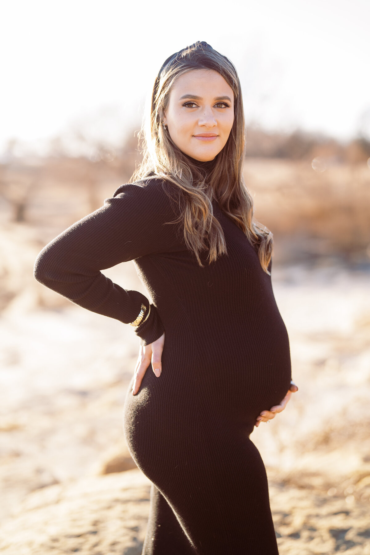 Portrait of a pregnant woman wearing black in a country field with one hand curled around her stomach on a sunny day.