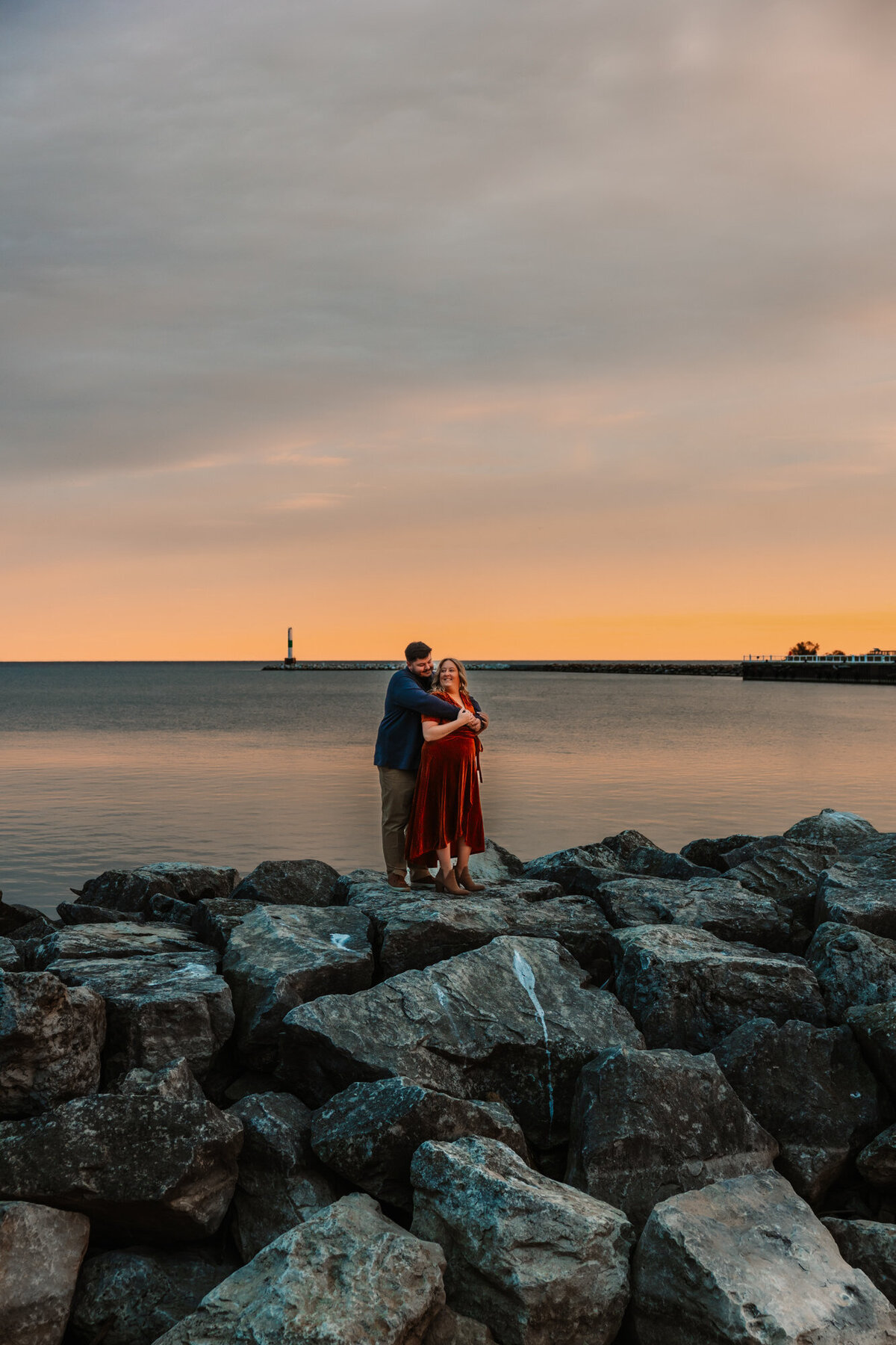 man holds woman from behind while they look at each other as the sun sets over lake michigan in port washington