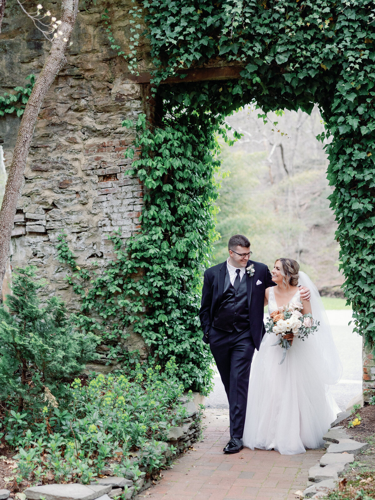 A bride and groom walk together under a stone archway that is covered in vibrant green ivy