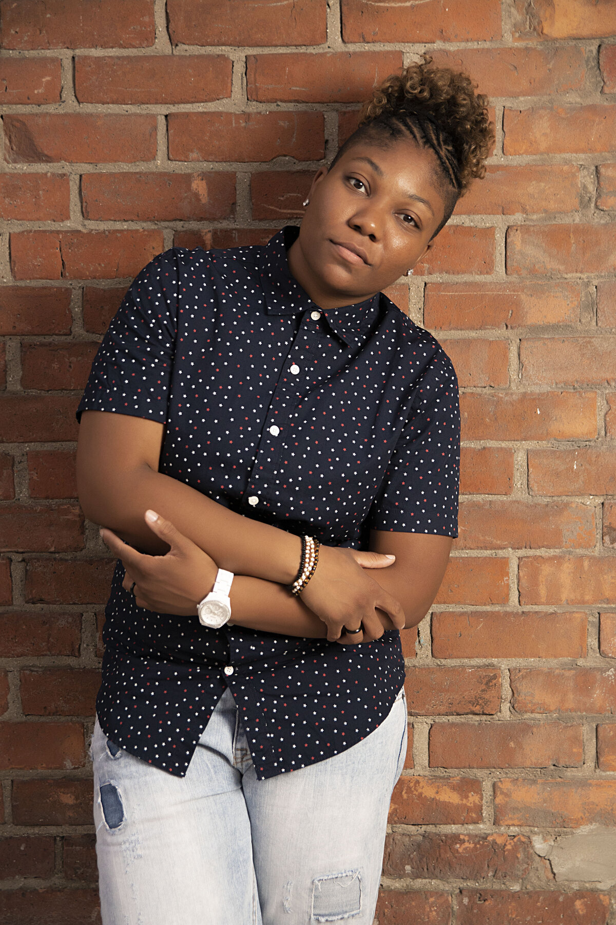 Woman in blue button up top, stonewash blue jeans and a watch standing in front of a brick wall