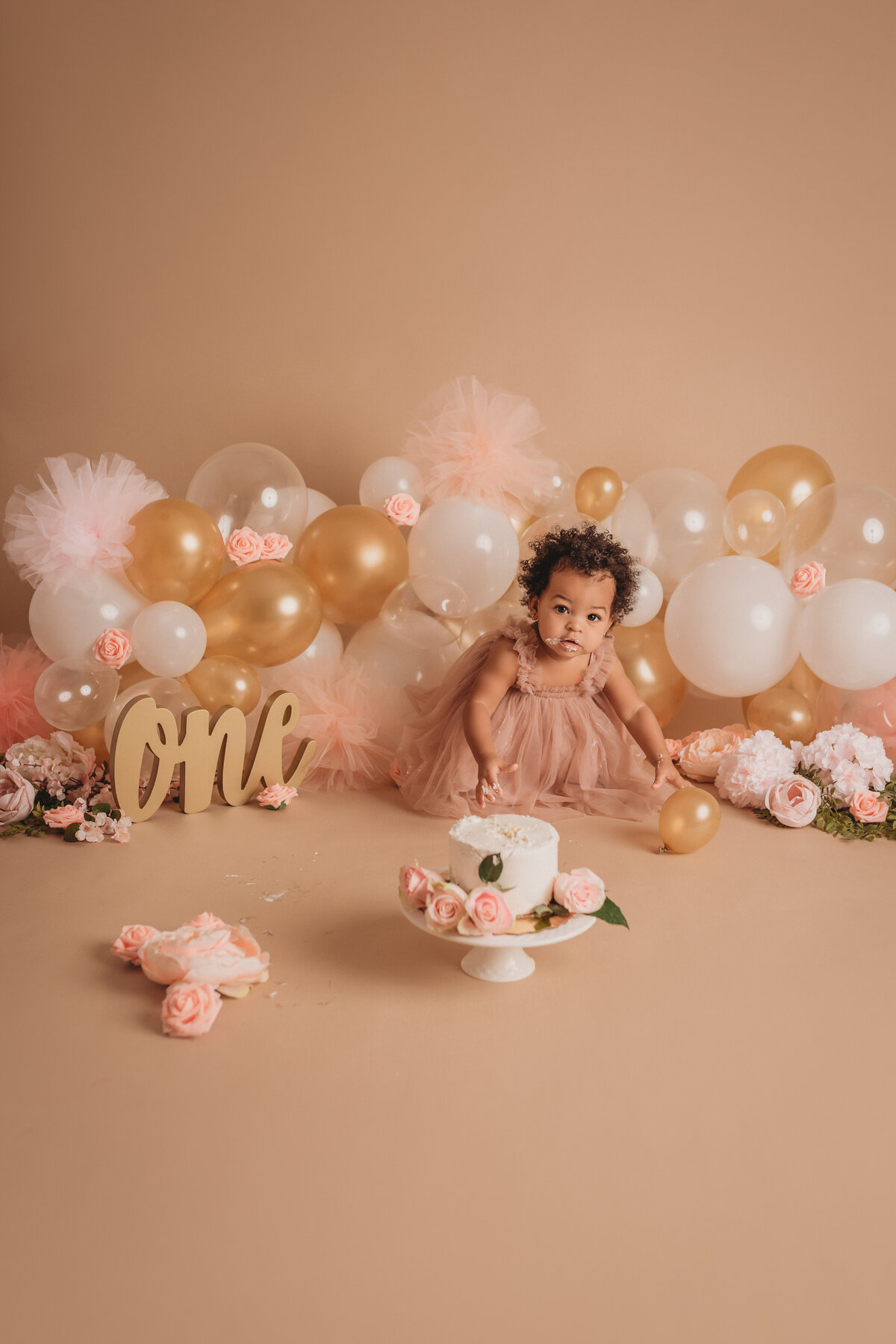 One year old baby girl in pink tulle dress on tan background with neutral colored balloon garland eating her white iced birthday cake with pink roses on it