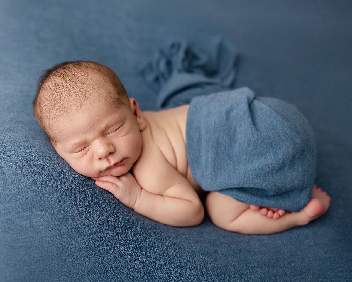 Newborn baby boy posed on a rich blue backdrop with a matching wrap across his waist, sleeping peacefully with hands under his cheek. Captured by Inland Empire newborn photographer Kaitlyn Dawn Photography.
