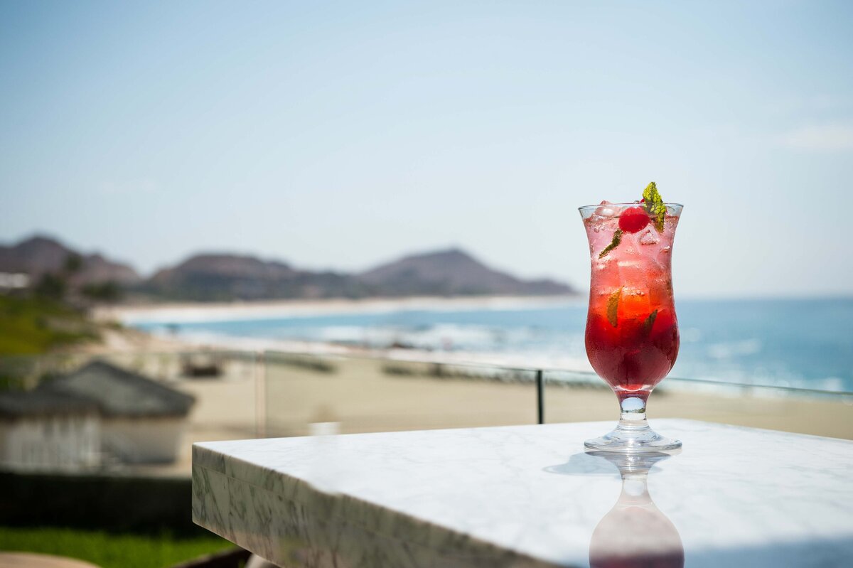 A red cocktail with condensation sits on bar counter with ocean view and beach in background