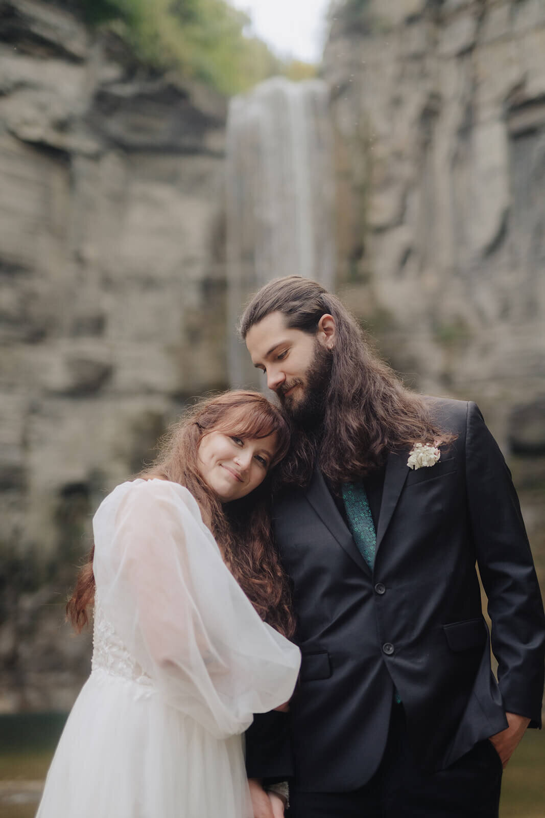 Bride resting head on grooms shoulder in front of a waterfall