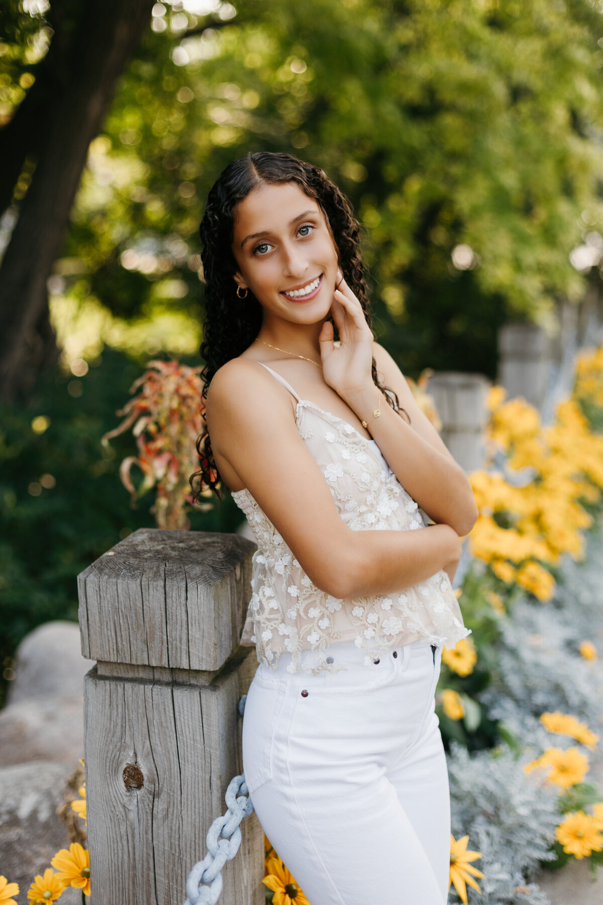 Senior girl in a white floral top and white pants leaning against a wooden pole in front of yellow flowers