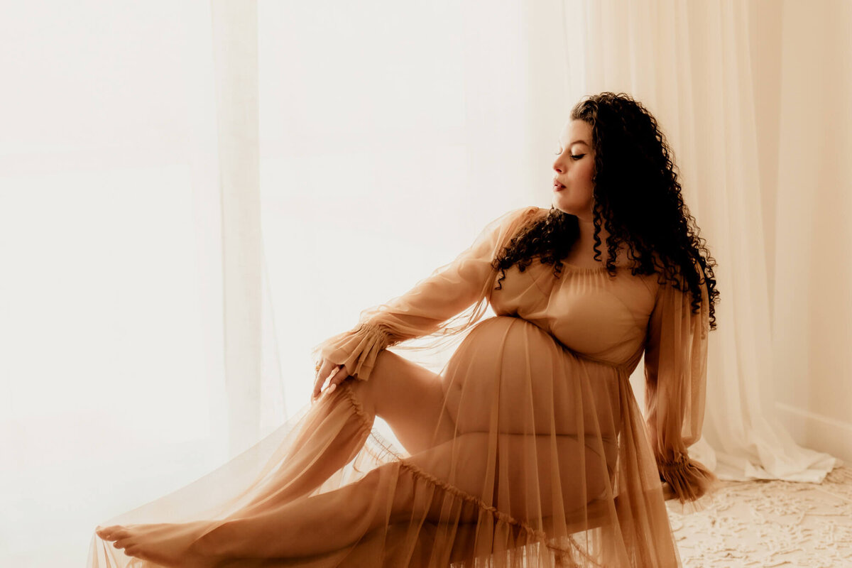 Pregnant woman with curly hair sitting on a bench her her wrist resting her her knee. 